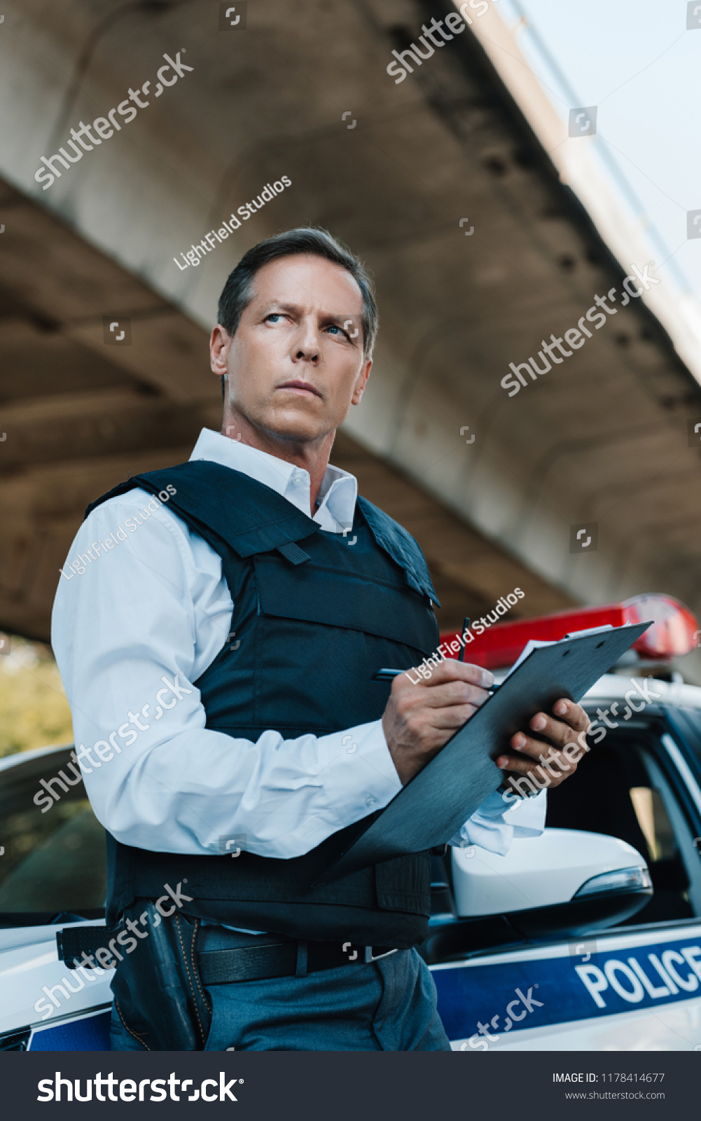 stock-photo-low-angle-view-of-middle-aged-male-police-officer-in-bulletproof-vest-writing-in-clipboard-near-car-1178414677